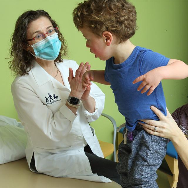 一个穿着蓝色衬衫、一头卷发的蹒跚学步的孩子被一位理疗师抱着，他举起双臂, as Stacy Suskauer look on. 